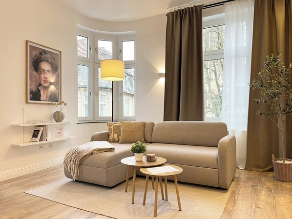 Een moderne woonkamer met een beige bank, een klein rond tafeltje, een vloerlamp en een groot raam met gordijnen.