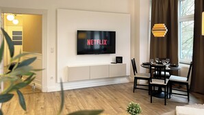 A modern living room with a mounted TV displaying "NETFLIX", a dining area with a table and chairs, and a large window with curtains.