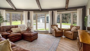 Ein geräumiges Wohnzimmer mit einem großen, braunen Ledersofa, zwei braunen Ledersesseln und einem Holztisch. Das Zimmer verfügt über große Fenster mit weißen Vorhängen, einen gemusterten Teppich und einen Holzboden.