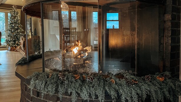 Ein gemütlicher Kamin mit Glaseinfassung, umgeben von immergrünen Zweigen und Tannenzapfen.