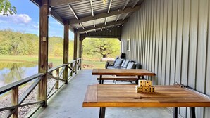 Ein überdachter Patio-Bereich mit Holztischen und -bänken, Blick auf einen Teich und eine Metallwand.