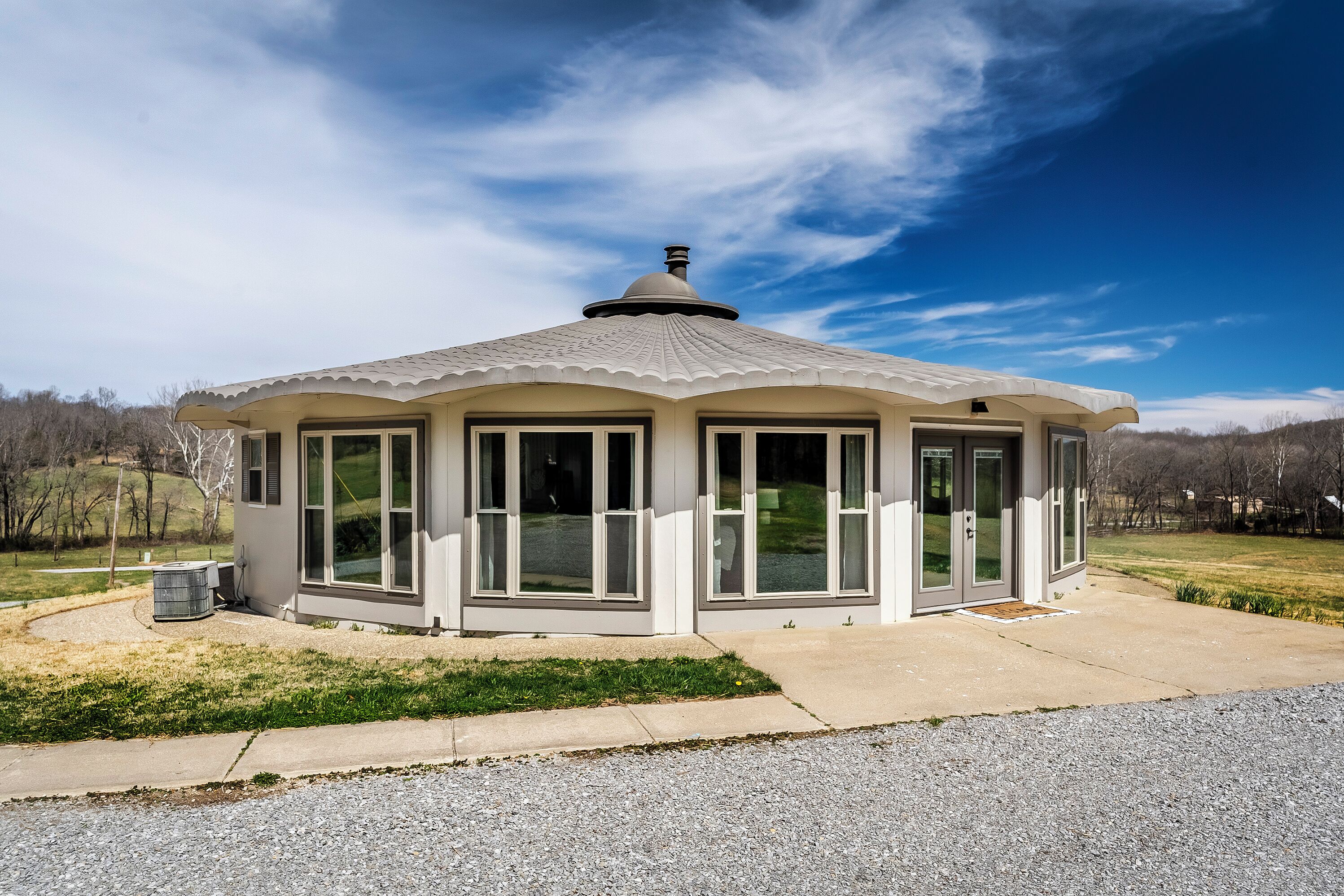 A small, white gazebo-style structure with large windows and a covered roof, surrounded by a grassy area and a gravel path.