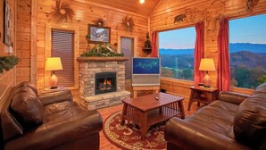 A cozy cabin interior with a stone fireplace, leather sofas, a TV, and a scenic mountain view.