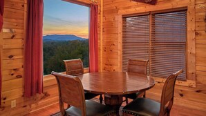A wooden cabin interior with a round dining table and four chairs, a window with blinds, and a view of a forested mountain range.