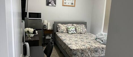 A compact hotel room with a bed, a desk, a chair, a microwave, and a small table with a vase of flowers.