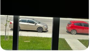 Une vue à travers une fenêtre, avec une voiture beige garée dans la rue et une voiture rouge garée sur le trottoir.