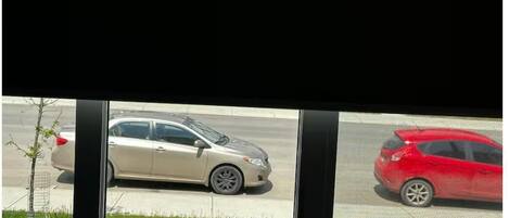 Vista a través de una ventana con un coche color beige estacionado en la calle y un coche rojo en la acera.