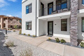 A modern two-story building with a stone accent wall, numbered 12, featuring a balcony with metal railings and a flat roof.
