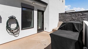 A modern outdoor patio with a covered grill, a stone wall, and a glass door leading to a covered entrance. - Touch of Class - 5BR - Private Hot Tub & Near Zion (Hurricane)