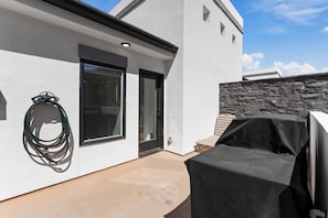 Een modern terras buiten met een overdekte barbecue, een stenen muur en een glazen deur naar een overdekte ingang.