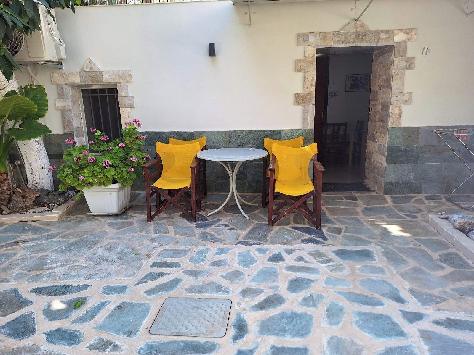 Une terrasse extérieure avec un sol en pierre, deux chaises en bois, une petite table ronde et un pot de fleurs orné de fleurs roses.