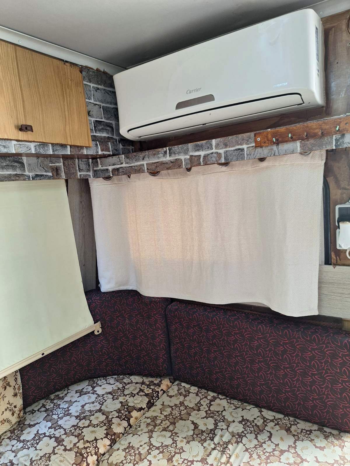 Ein kompaktes Wohnmobil-Interieur mit einer fest installierten Klimaanlage, einem Holzschrank, einer gepolsterten Sitzbank und einer geblümten Tagesdecke.
