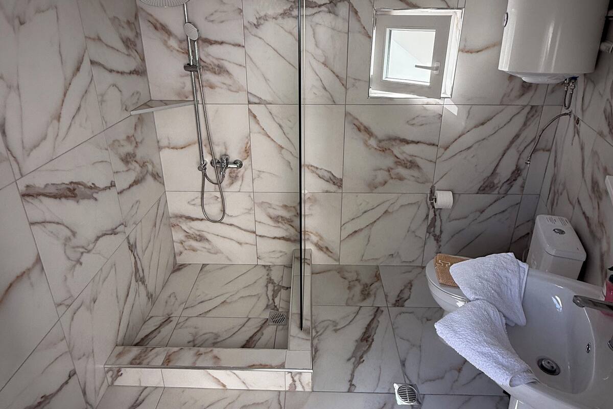 Baño con azulejos de mármol, ducha con cerramiento de vidrio, lavamanos blanco y perchero.