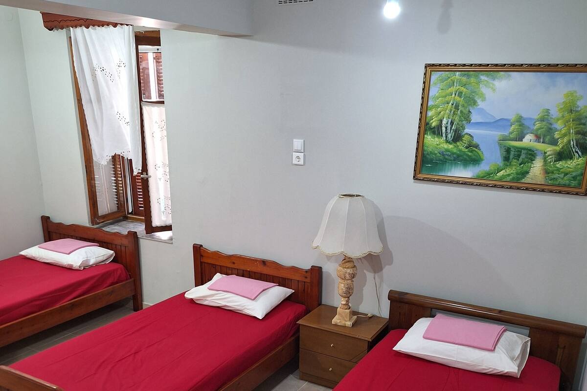 Una habitación con tres camas de madera, cada una con una colcha roja y almohadas blancas. Hay una mesita de noche de madera con una lámpara entre las camas. Un cuadro de un paisaje cuelga en la pared.