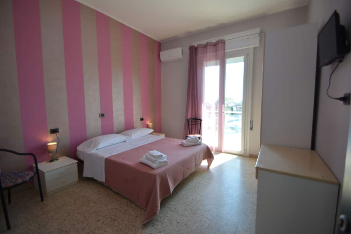 Un dormitorio con cama, mesitas de noche, una silla, televisor y ventana con cortinas.