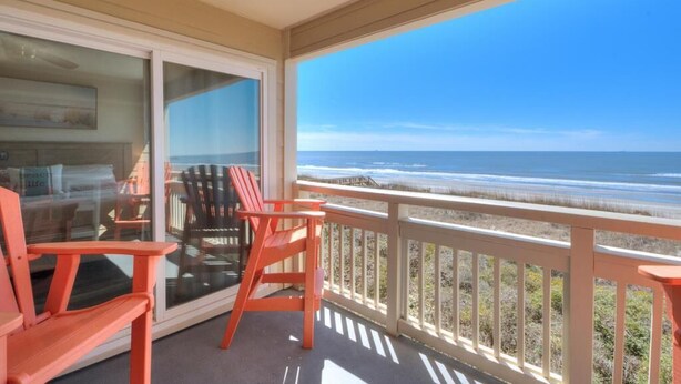 Een balkon met uitzicht op zee, twee rode stoelen en een glazen deur die leidt naar een kamer met een bed en een schilderij.