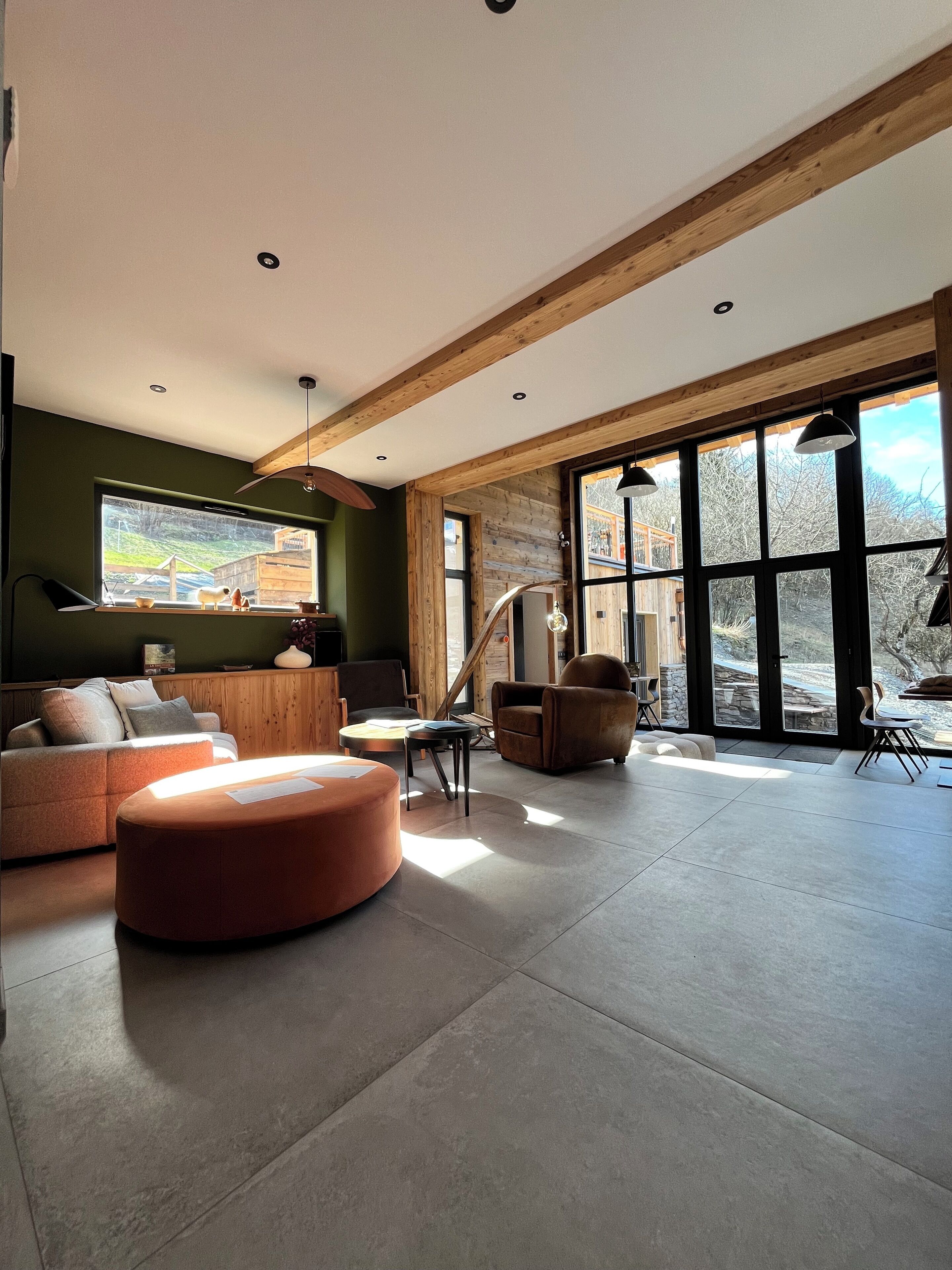 A spacious living area with a large window, wooden beams, a sofa, a round ottoman, and a chair.
