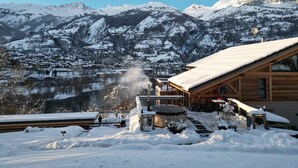 A wooden cabin with a snow-covered roof, a hot tub, and a view of a mountain town and snow-covered mountains in the background. - La Grange des Eulets (Bourg-Saint-Maurice)