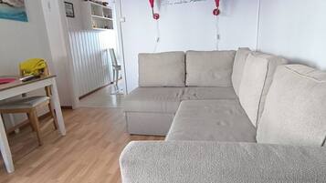 Un salon avec un canapé d’angle beige, une table en bois recouverte d’une nappe jaune et un mur blanc orné d’une étagère rouge où sont rangés des livres.