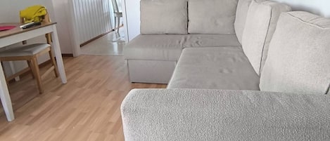 Ein Wohnzimmer mit einem beigefarbenen Ecksofa, einem Holztisch mit gelber Tischdecke und einer weißen Wand mit rotem Regal, das Bücher beherbergt.