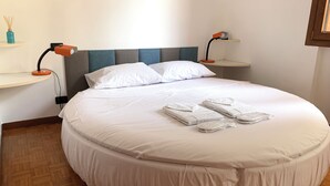 Una cama bien hecha con ropa blanca y toallas, un cabecero gris y azul, mesitas de noche con lámparas, y un pequeño estante con un objeto decorativo.