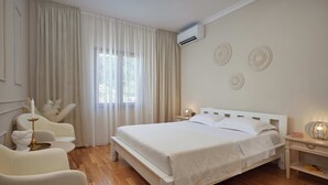 Una camera da letto con un letto bianco, un comodino con una lampada, una sedia e una finestra con tende.