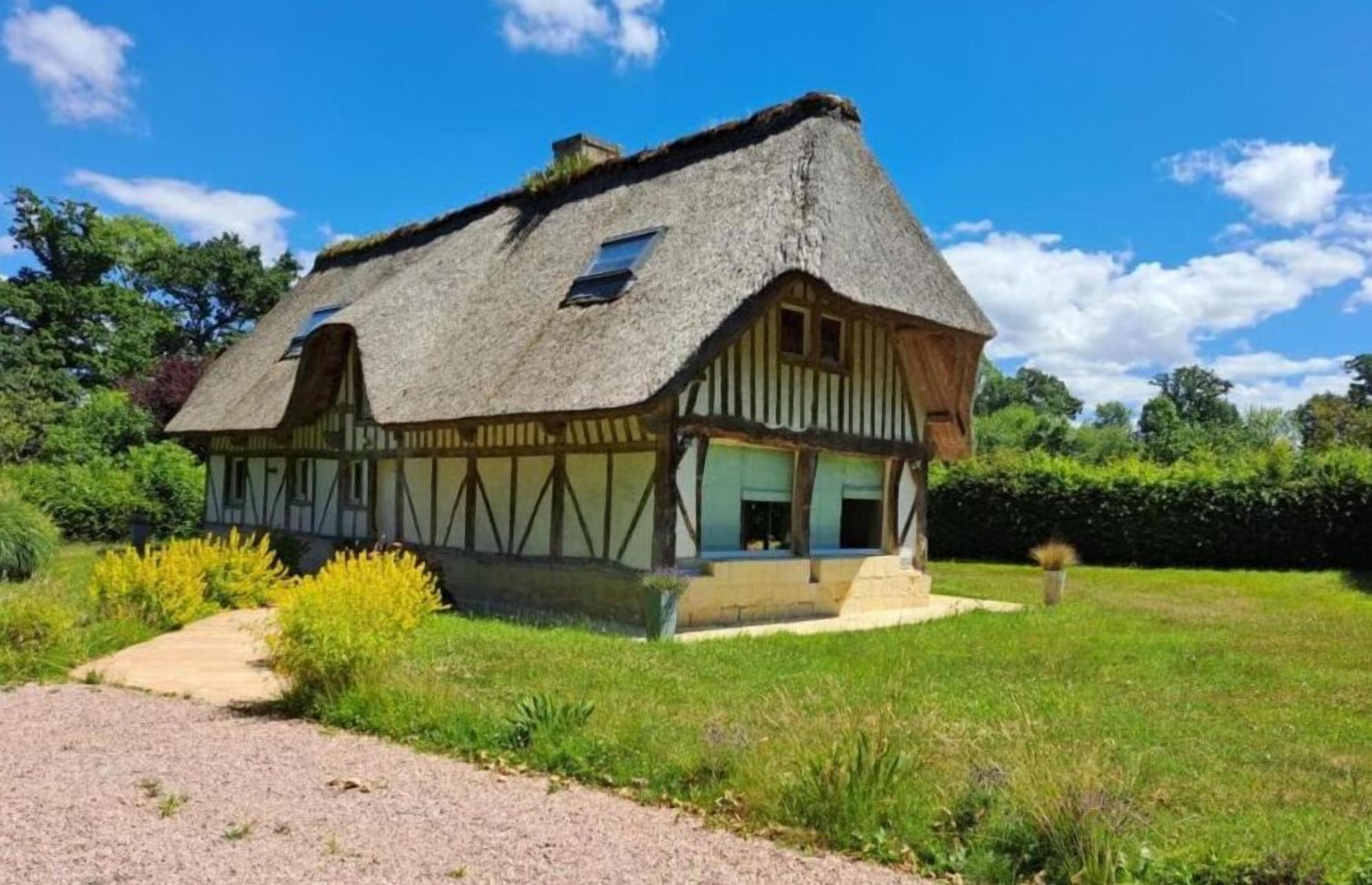 Une maison traditionnelle à colombages, dotée d’un toit de chaume, entourée d’un jardin bien entretenu et d’un chemin de gravier menant à l’entrée.
