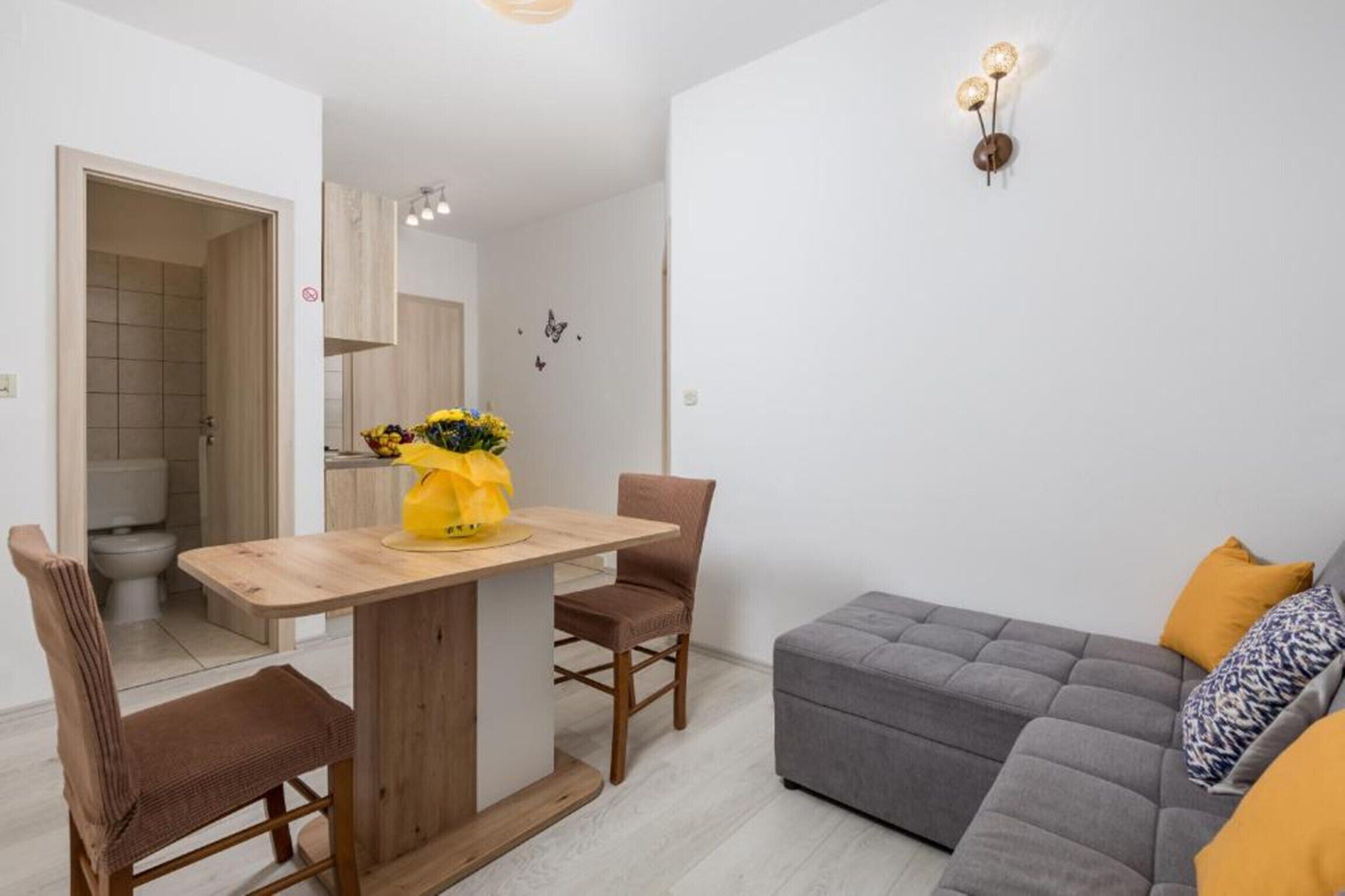 A modern living room with a grey sofa, a wooden dining table with two chairs, and a small kitchen area with a white cabinet.