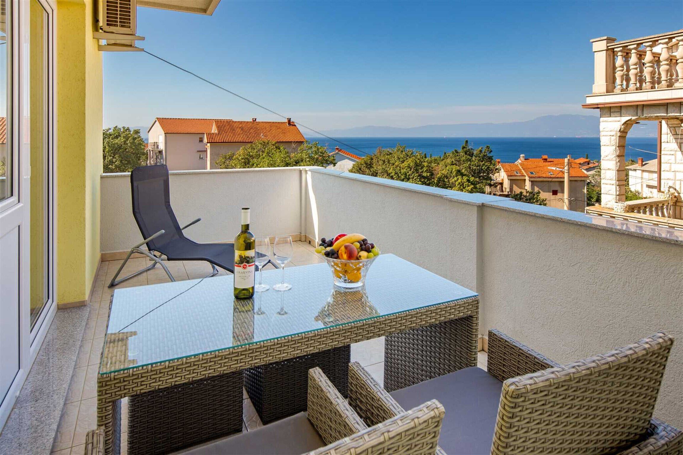 A balcony with a glass table, wicker chairs, a bottle of wine, and a bowl of fruit.
