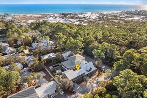 An aerial view of a residential area with houses, dense greenery, and a beach in the background.
