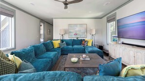 A living room with a sectional sofa, a coffee table, a ceiling fan, a large TV, and a painting on the wall. - Newly Built Lux Grayton Beach Home "Timeless 30A" Pool, Golf Cart, Bikes (Santa Rosa Beach)