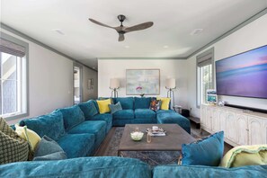A living room with a sectional sofa, a coffee table, a ceiling fan, a large TV, and a painting on the wall. - Newly Built Lux Grayton Beach Home "Timeless 30A" Pool, Golf Cart, Bikes (Santa Rosa Beach)