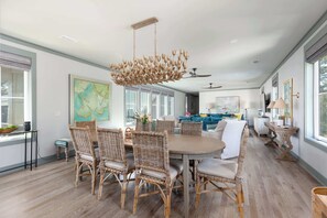 A spacious dining area with a round table and wicker chairs, a chandelier, and large windows providing natural light. - Newly Built Lux Grayton Beach Home "Timeless 30A" Pool, Golf Cart, Bikes (Santa Rosa Beach)