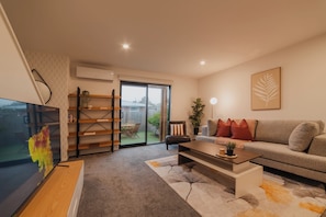 A modern living room with a sofa, coffee table, and TV. There is a bookshelf, a plant, and a window with a view.