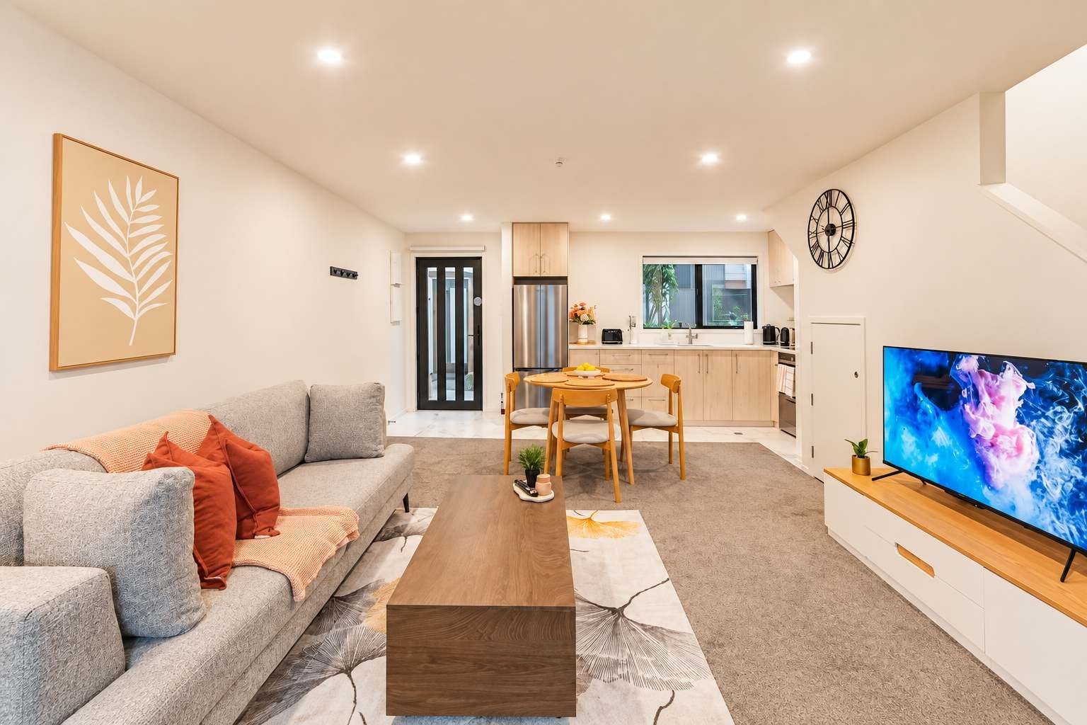 A modern living room with a sofa, coffee table, and a TV. There is a dining area with a table and chairs. The kitchen is visible in the background with cabinets and a window.