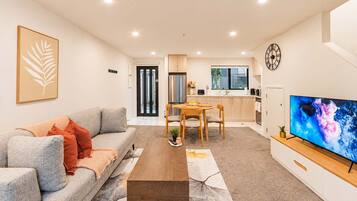 A modern living room with a sofa, coffee table, and a TV. There is a dining area with a table and chairs. The kitchen is visible in the background with cabinets and a window.