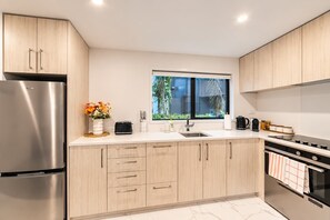 A modern kitchen with stainless steel appliances, light wood cabinetry, a window with a view, and a marble countertop.