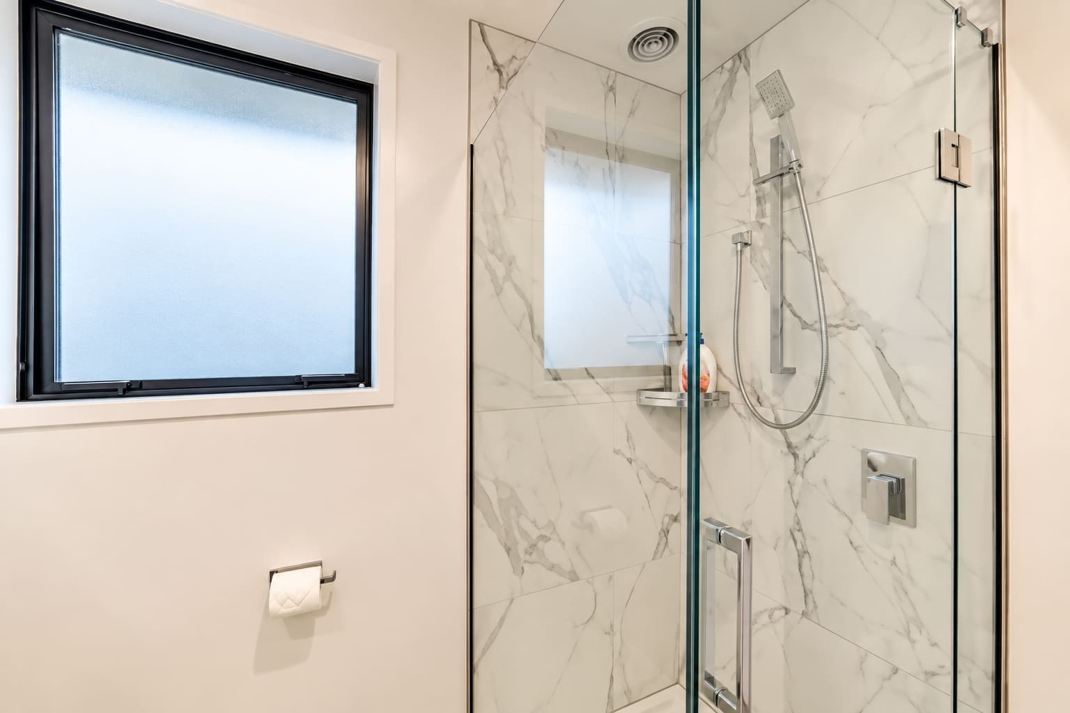 Un baño moderno con cabina de ducha de vidrio, un dispensador de papel higiénico y un ventanal.