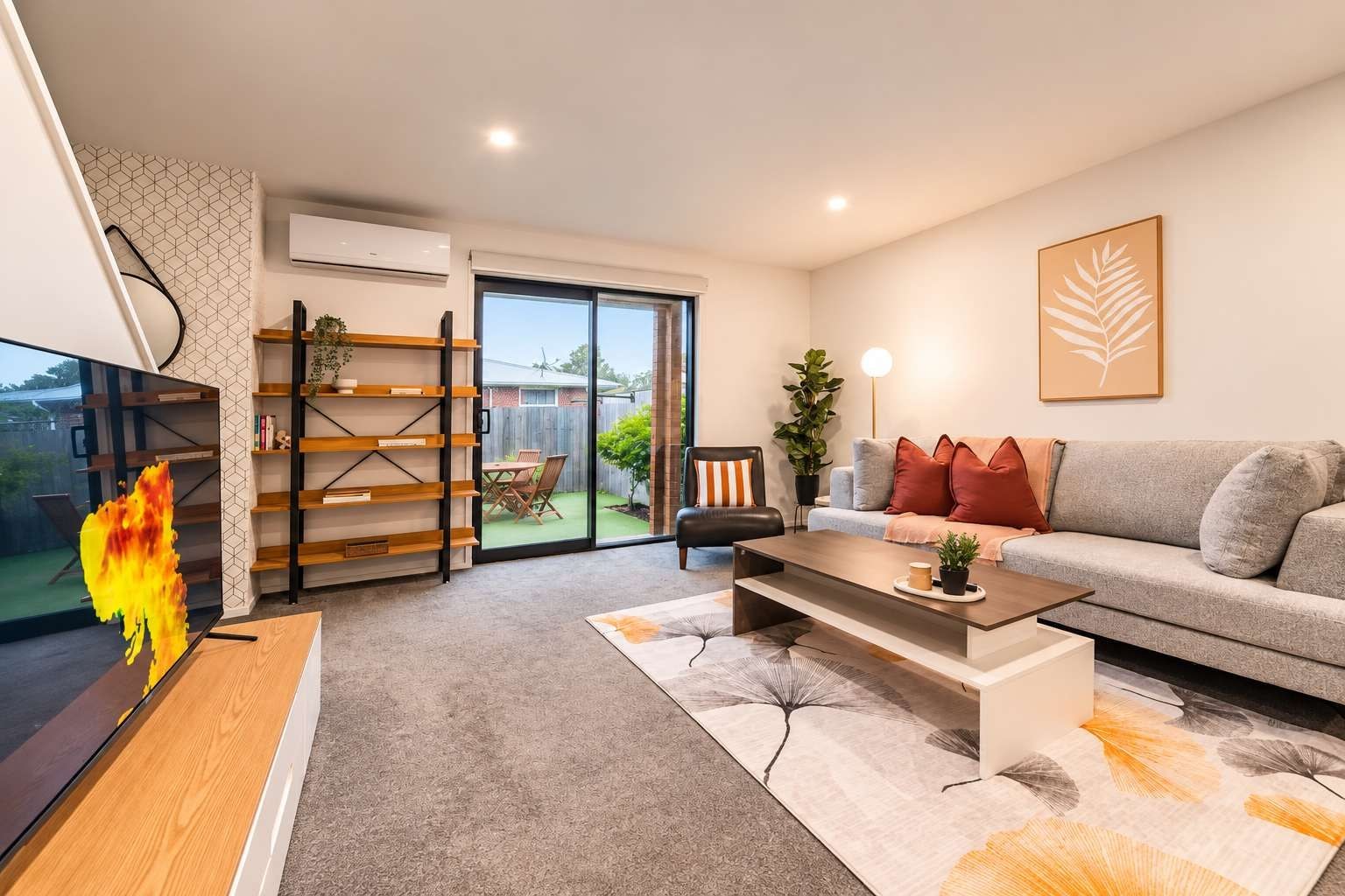 A modern living room with a sofa, coffee table, and TV. There is a bookshelf, a plant, and a window with a view of the outdoors.