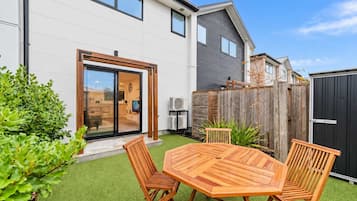 Una casa moderna con un patio, una mesa de madera con sillas y un jardín trasero cercado.