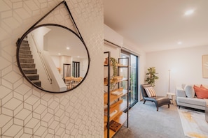 A modern interior with a geometric patterned wall, a round mirror, a staircase, a dining area, a living room with a sofa and a coffee table, and a bookshelf.