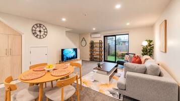 A modern living room with a dining area, a grey sofa, a TV, and a bookshelf.