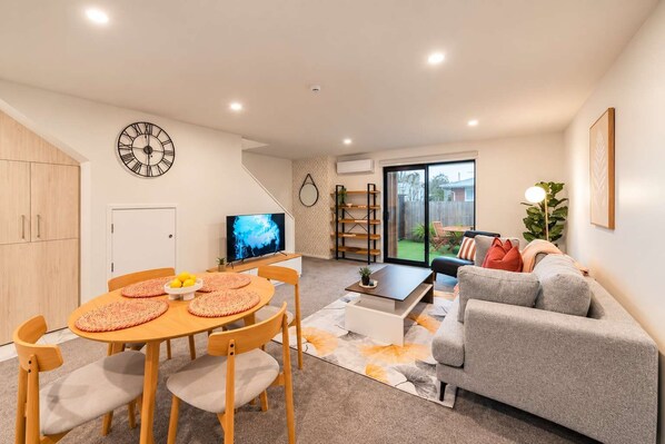 A modern living room with a dining area, a grey sofa, a TV, and a bookshelf.