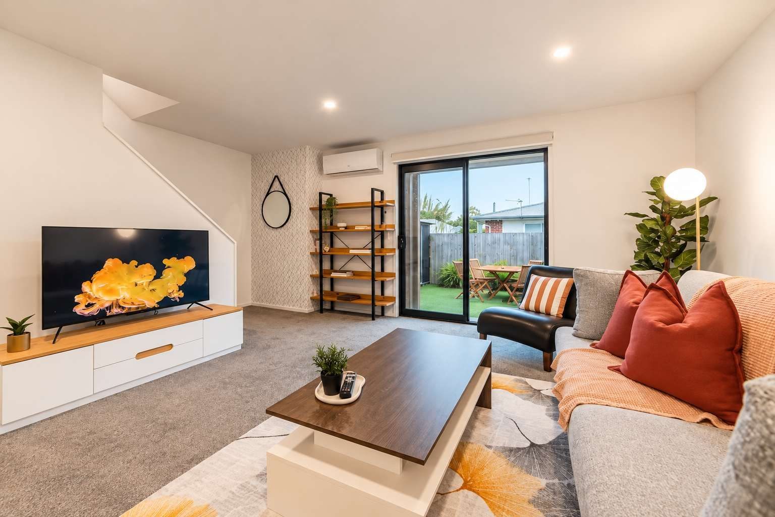 Un salón moderno con televisor de pantalla plana, una mesa de centro de madera, un sofá con cojines, una estantería y un ventanal corredero que da a un patio exterior.