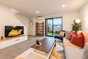 A modern living room with a flat-screen TV, a wooden coffee table, a sofa with cushions, a bookshelf, and a sliding glass door leading to an outdoor patio.