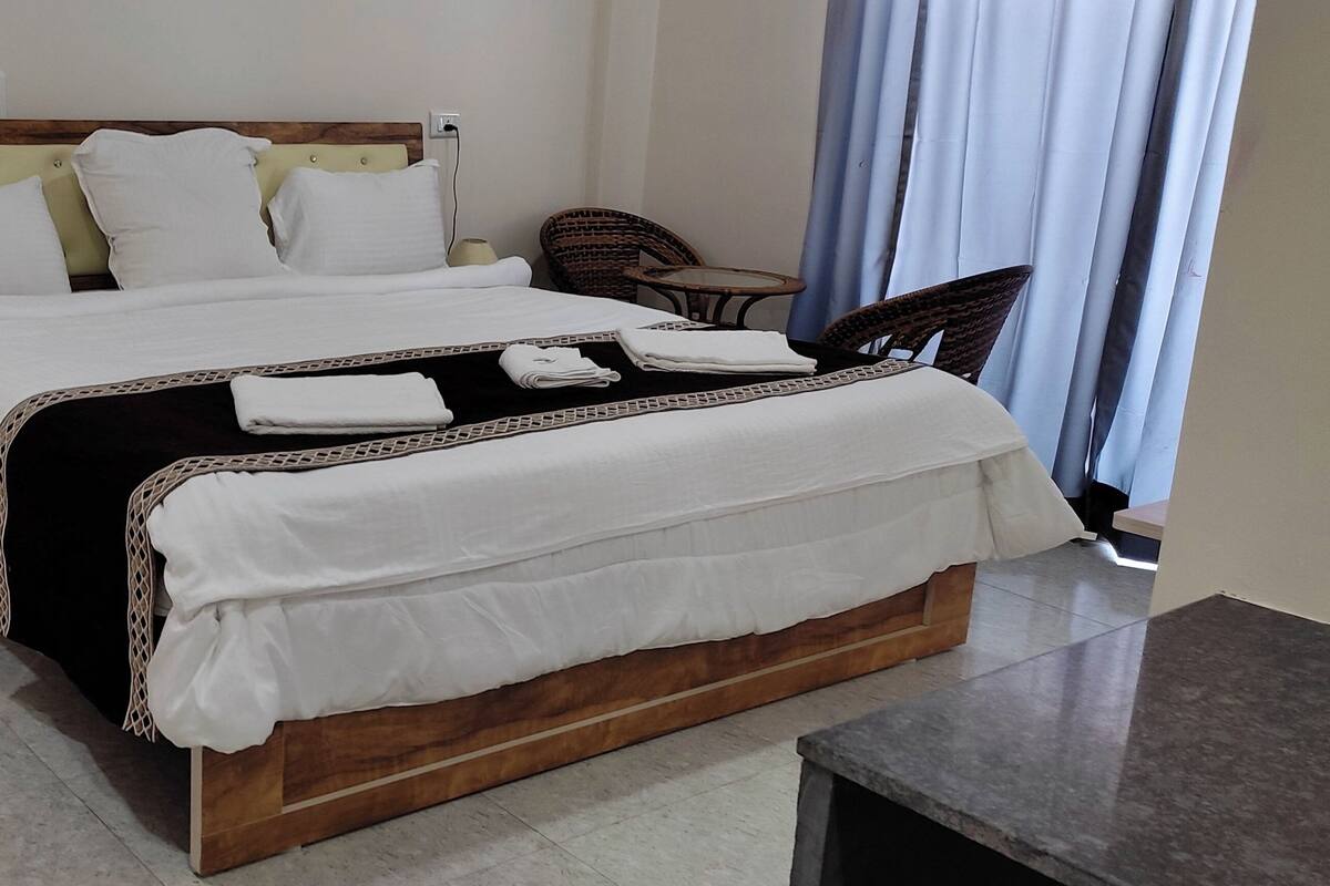 Un lit bien fait, avec du linge de lit blanc et un couvre-lit noir, une table de chevet en bois avec un tiroir, une chaise en osier et une petite table ronde avec une lampe.