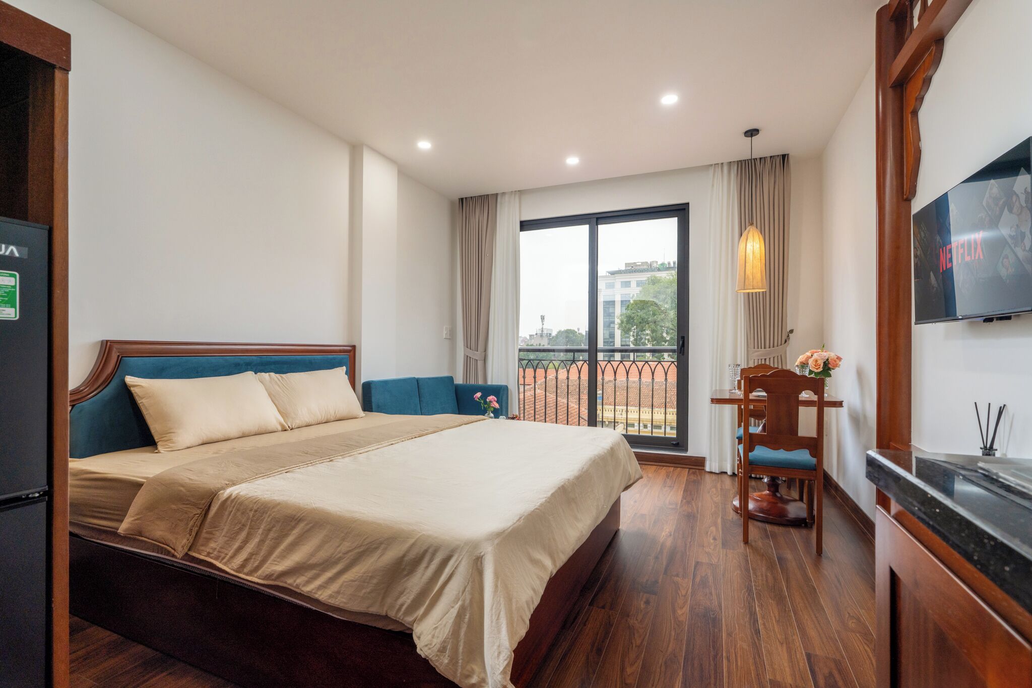 A hotel room with a large bed, a TV, a dining table with chairs, and a balcony with a view of the city.
