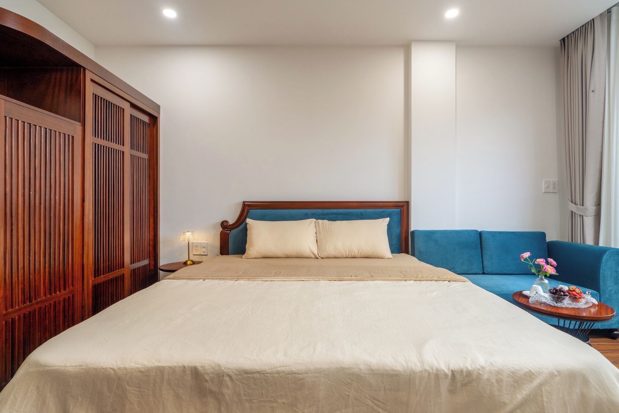 A hotel room with a large bed, a blue sofa, a wooden wardrobe, and a nightstand with a lamp.