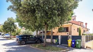 Una calle residencial con un SUV estacionado, una acera bordeada de árboles y una casa con entrada protegida por un muro.