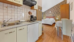 Cocina compacta con un lavamanos, cafetera y un refrigerador pequeño. Se ve una cama con cabecera blanca al fondo.